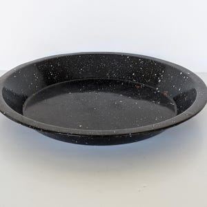 Könnte beinhalten: Eine schwarze Emaille-Kuchenplatte mit weißen Sprenkeln. Die runde Platte hat einen leicht erhöhten Rand und eine glänzende Oberfläche. Dieses Vintage-Küchengeschirr eignet sich zum Backen und Servieren.