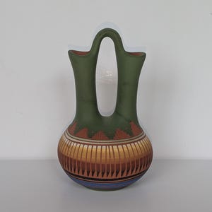 May include: A double-spouted ceramic vase featuring a matte olive green upper section and a rounded base decorated with horizontal bands of terracotta, yellow, and blue. The vessel has a central arched handle connecting the two vertical spouts.