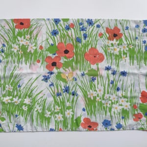 Puede incluir: Una tela rectangular blanca con un estampado floral. El diseño presenta amapolas rojas, flores silvestres azules y margaritas blancas en medio de hierba verde. También se ve una mariposa amarilla.