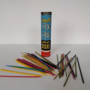 May include: A vintage tin of "Plastic Pix Pick-Up Sticks" with a blue background and yellow lettering. The tin contains 31 plastic sticks in various colors, including red, yellow, blue, green, and brown.