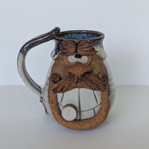 May include: A ceramic mug featuring a sculpted face with a prominent brown mustache, bushy eyebrows, and a wide grin showing teeth. The mug has a textured, earthy brown finish on the face and a smooth, mottled blue and white glaze on the rim.