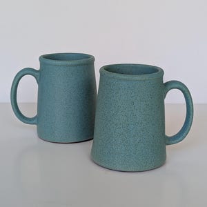 May include: Two matching mugs with a speckled teal finish and a cylindrical shape. Each mug features a sturdy handle on the side and a smooth, rounded rim. The surface has a consistent matte texture with small dark flecks throughout the design.