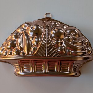 May include: A copper-colored metal mold shaped like a basket overflowing with fruit. The mold has a detailed design with a handle at the top for hanging. The basket design includes a woven pattern and a decorative border.