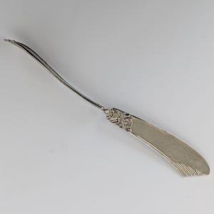Può includere: Un coltello da burro argentato con manico curvo e lama larga e piatta. Il manico presenta dettagli ornati incisi. La lama ha una forma leggermente curva e un bordo strutturato. Il coltello è posto su uno sfondo chiaro e semplice.