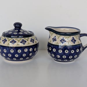 Pode incluir: Uma açucareiro de cerâmica com tampa e um jarro de creme a condizer. Ambas as peças são decoradas com um padrão floral azul e branco com uma borda azul escura. O açucareiro é redondo e o jarro tem uma pega e um bico.