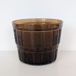 May include: A brown glass planter with a barrel-like design. The planter has a textured, vertical wood grain pattern and is encircled by two horizontal bands. The top rim is a lighter shade of brown.