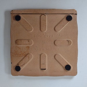 May include: A square, light brown ceramic tile with a textured surface. The tile has black rubber feet and the text "schingies" and "MADE IN ITALY" printed on it.