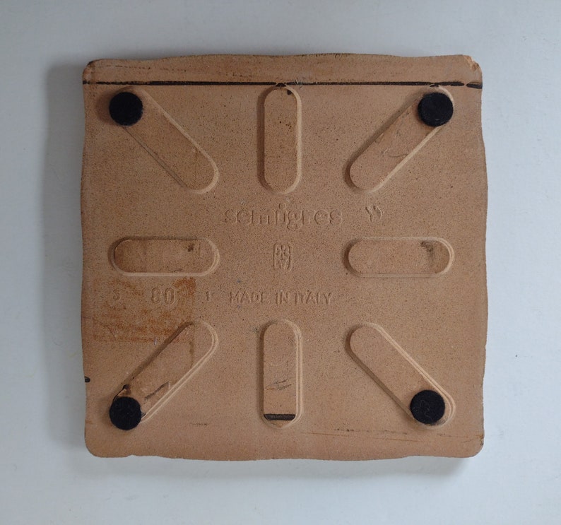 May include: A square, light brown ceramic tile with a textured surface. The tile has black rubber feet and the text "schingies" and "MADE IN ITALY" printed on it.