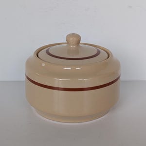 May include: A beige ceramic sugar bowl with a lid. The bowl has a rounded shape with a dark brown stripe around the middle and a matching stripe on the lid. The lid has a small knob on top.