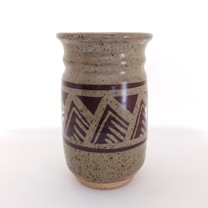 May include: A speckled, tan-colored ceramic vase with a dark brown geometric pattern. The vase has a cylindrical shape with a slightly flared rim and a textured surface. The design features repeating triangles.