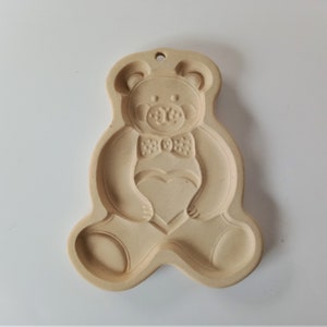 May include: A beige ceramic cookie cutter shaped like a teddy bear holding a heart. The bear is wearing a bow tie.