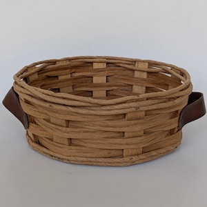Könnte beinhalten: Ein runder, geflochtener Korb in Naturtönen. Er hat zwei dunkelbraune Ledergriffe. Der Korb besteht aus geflochtenen Materialstreifen und wirkt rustikal.