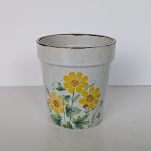 May include: A ceramic flower pot with a speckled, light gray finish. The pot features a hand-painted design of yellow daisies with green leaves and stems. The rim of the pot has a darker brown trim.