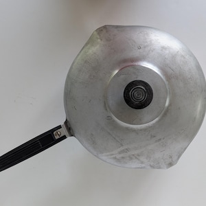 May include: Overhead view of a vintage aluminum saucepan with a black handle. The pan has a circular body with a spout and a black knob in the center. The handle is attached with a metal bracket.
