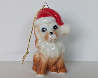 May include: A ceramic dog ornament wearing a red and white Santa hat. The dog has brown and white fur, black eyes, and a gold string for hanging. This festive decoration is perfect for Christmas.