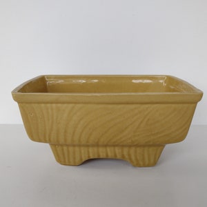 以下が含まれることがあります： 温かみのあるマスタードイエローの正方形のセラミックプランター。側面には木目調のテクスチャがあり、3つの小さな一体型脚の上に置かれています。内部は滑らかです。シンプルなデザインで、さまざまな植物に適しています。寸法はcmです。