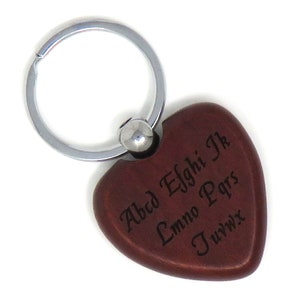 May include: A brown wooden heart-shaped keychain with a silver metal ring. The heart has the letters of the alphabet engraved on it in a cursive font.