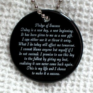 May include: Black round pendant with a silver chain. The pendant has white text that reads "Pledge of Success" and includes a motivational quote about making the most of each day.