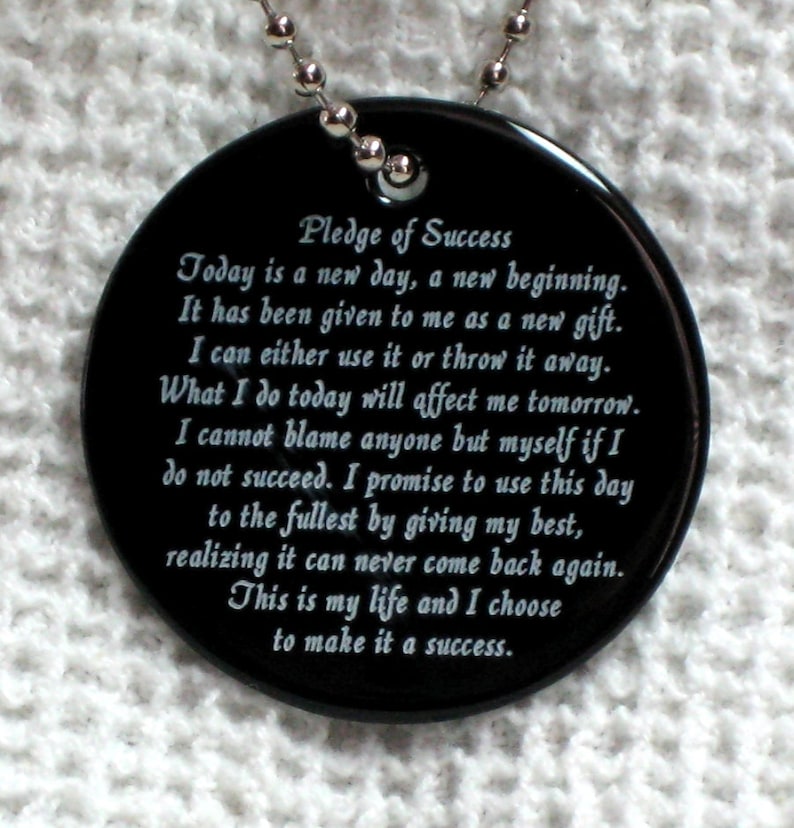 May include: Black round pendant with a silver chain. The pendant has white text that reads "Pledge of Success" and includes a motivational quote about making the most of each day.