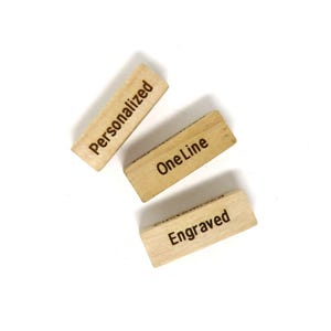 May include: Three small, rectangular wooden blocks with engraved text. One block reads "Personalized", another "One Line", and the third "Engraved". The blocks are light brown and arranged on a white background.