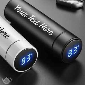 May include: Two insulated water bottles, one black and one white, with digital temperature displays showing 83°C. The black bottle has the text "Your Text Here" in white script. The white bottle has the text "Here" in white script.