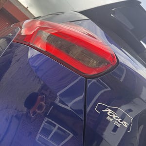 Może przedstawiać: Zbliżenie na tył niebieskiego samochodu z czerwonym światłem tylnym. Biała naklejka z napisem "Focus" i latami "1998-2025" jest widoczna na karoserii. Powierzchnia samochodu odbija otoczenie.