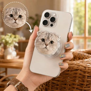 May include: A light-colored smartphone featuring a circular, collapsible phone grip attached to the back. The grip displays a high-resolution, detailed portrait of a grey and white Scottish Fold cat with large, expressive eyes and folded ears.