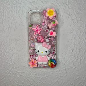 May include: Clear phone case decorated with pink and white embellishments. Features a Hello Kitty figure, a pink star, flowers, and a rainbow-colored swirl. The case is filled with small pink beads.