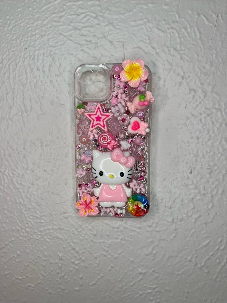 May include: Clear phone case decorated with pink and white embellishments. Features a Hello Kitty figure, a pink star, flowers, and a rainbow-colored swirl. The case is filled with small pink beads.