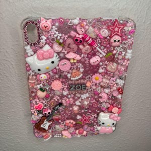 May include: A clear phone case adorned with an array of pink and white embellishments. Features include Hello Kitty figures, stars, hearts, flowers, and beads. The case also has a small guitar and the name "ZOE" in silver letters.