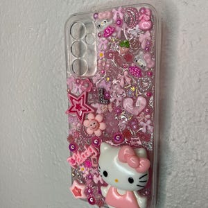 May include: Clear phone case decorated with pink embellishments, including stars, flowers, bows, and a Hello Kitty figure. The word "melody" is also visible. The case is designed to fit a phone with three camera lenses.