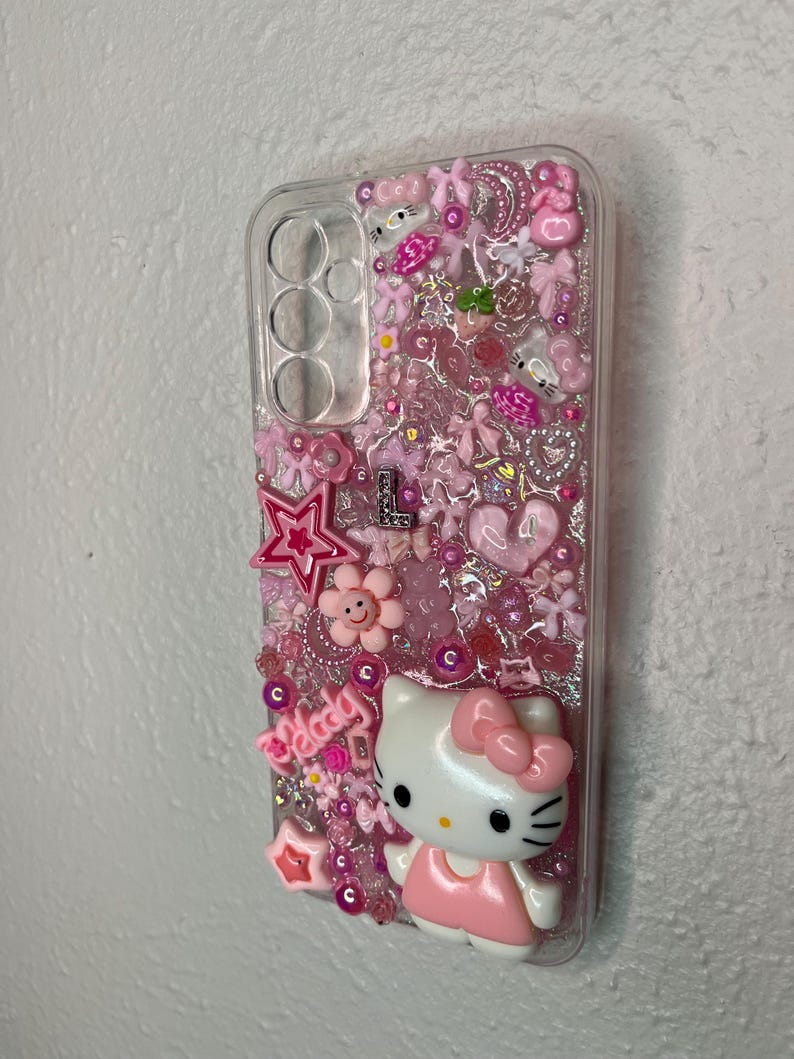 May include: Clear phone case decorated with pink embellishments, including stars, flowers, bows, and a Hello Kitty figure. The word "melody" is also visible. The case is designed to fit a phone with three camera lenses.