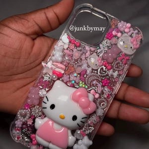 May include: Clear phone case decorated with pink and white Hello Kitty figures, bows, and other embellishments. The case has a 3D effect with various charms and glitter. The text "@junkbymay" is visible on the case.