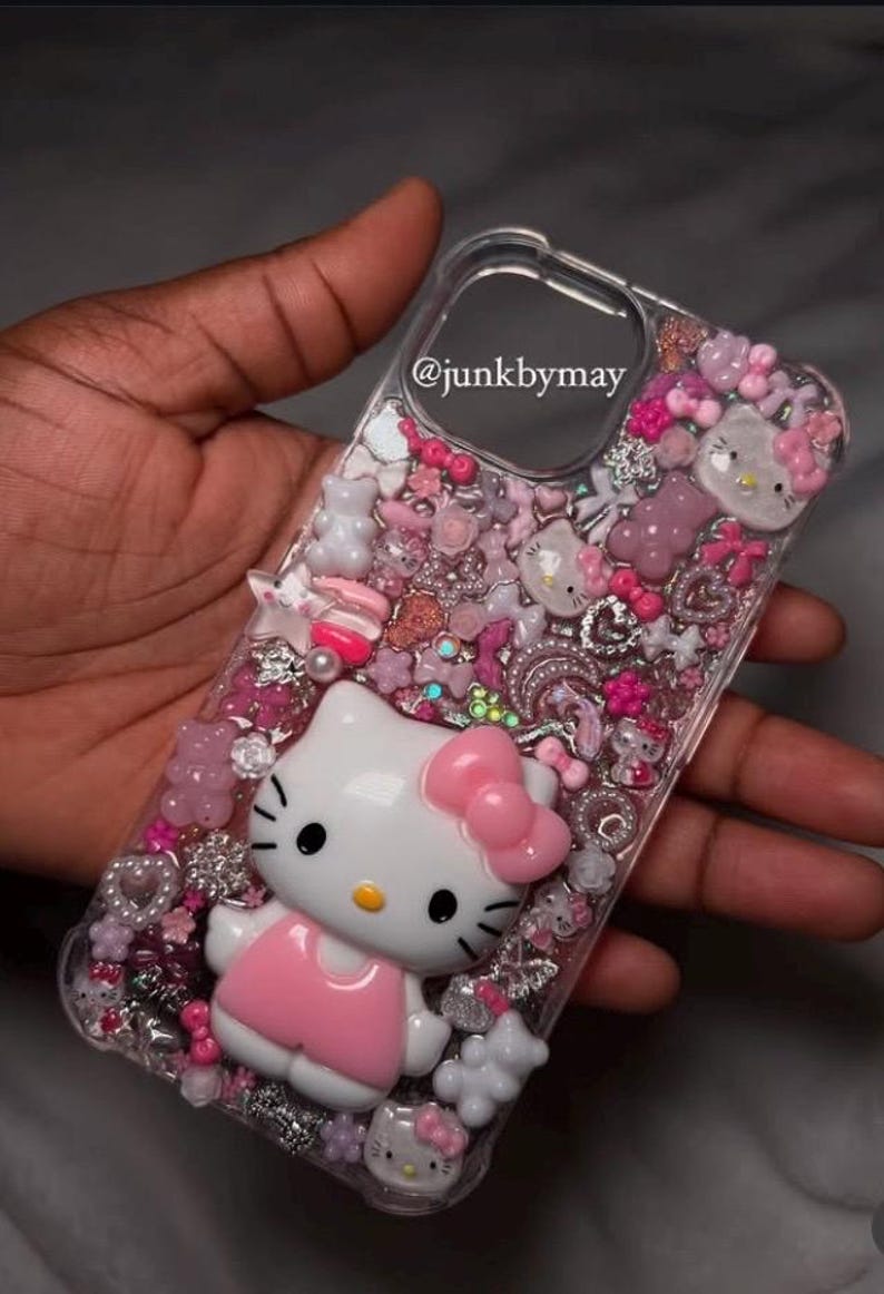 May include: Clear phone case decorated with pink and white Hello Kitty figures, bows, and other embellishments. The case has a 3D effect with various charms and glitter. The text "@junkbymay" is visible on the case.