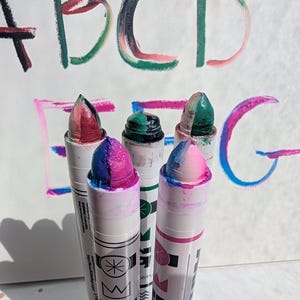 May include: Close-up of five colorful art crayons with a white background. The crayons are multi-colored with pink, blue, green, and red tips. The letters A, B, C, D, E, F, and G are painted in the background with the same colors.