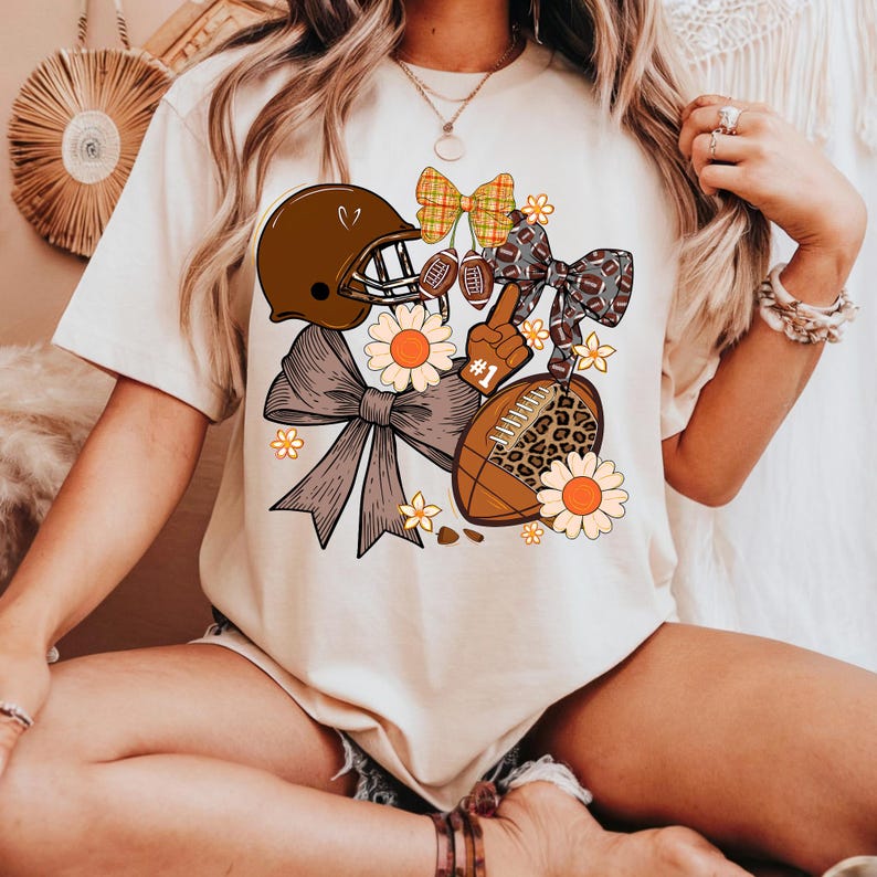 May include: Cream-colored t-shirt featuring a football-themed graphic. The design includes a brown football helmet, bows, a football, and daisies. The football has a leopard print pattern. The graphic also includes a foam finger with the number one.