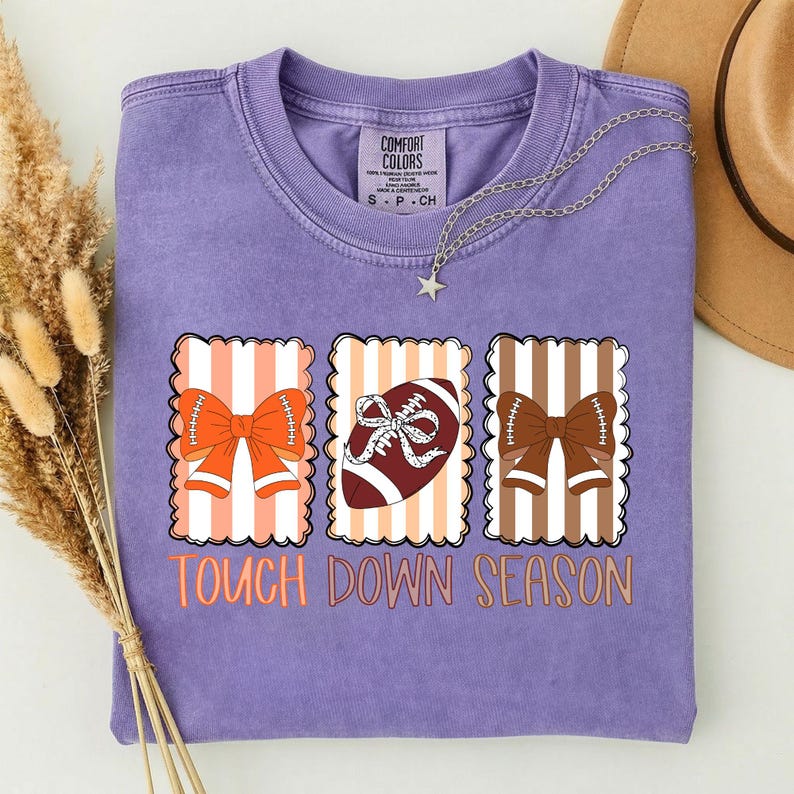 May include: A purple Comfort Colors t-shirt with a football theme. The shirt features three framed designs with bows and a football, and the words "TOUCH DOWN SEASON" in orange. A silver necklace with a star pendant is draped on the shirt.