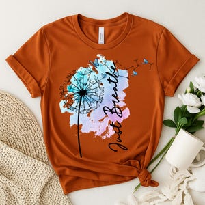 May include: Burnt orange t-shirt featuring a dandelion design with the words "Just Breathe" in a script font. The dandelion is in shades of blue, pink, and purple, with butterflies. The shirt is knotted at the bottom.