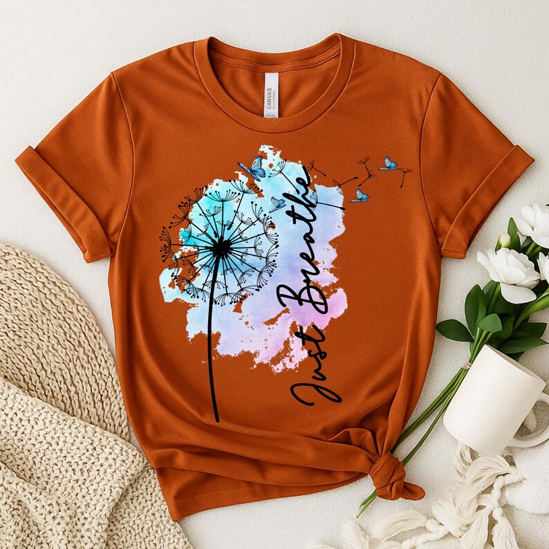 May include: Burnt orange t-shirt featuring a dandelion design with the words "Just Breathe" in a script font. The dandelion is in shades of blue, pink, and purple, with butterflies. The shirt is knotted at the bottom.