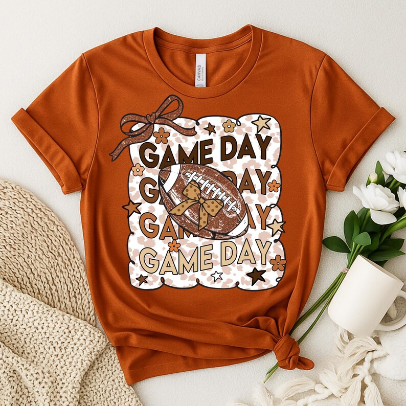 May include: Orange t-shirt with a football graphic and the words "Game Day." The graphic includes a football with a bow, stars, and flowers. The shirt is tied at the bottom.