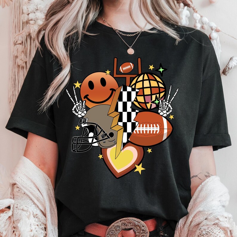 May include: Black t-shirt featuring a colorful graphic design. The design includes a football, a football helmet, a smiley face, a disco ball, a lightning bolt, a heart, and peace signs. The design is accented with yellow stars.