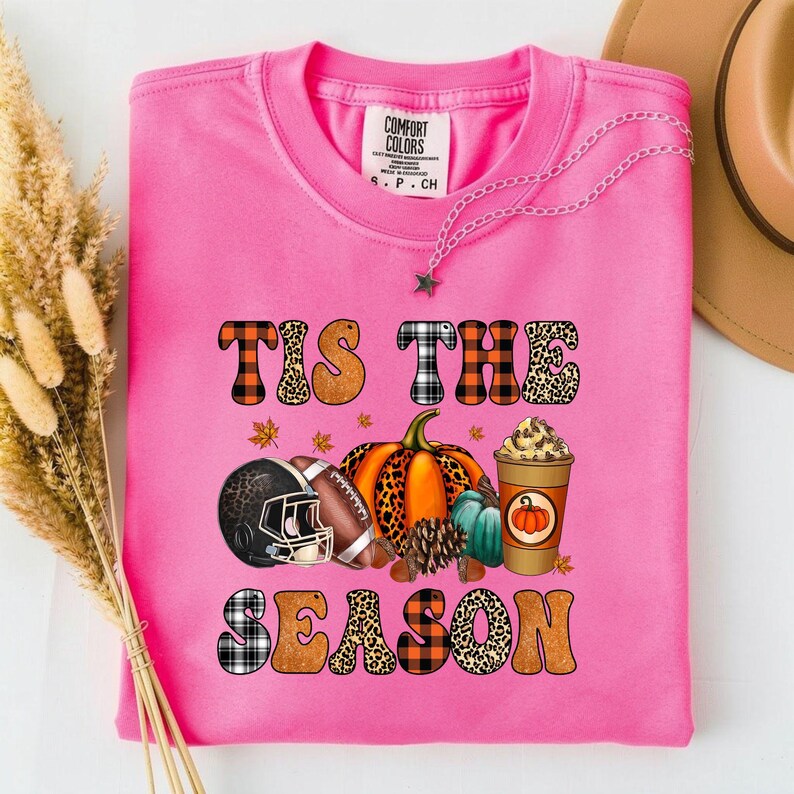 May include: A bright pink t-shirt with the phrase "Tis the Season" in a mix of plaid, leopard print, and orange lettering. The graphic includes a football helmet, a football, pumpkins, and a coffee cup.