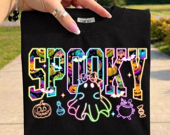 May include: A black t-shirt featuring the word SPOOKY in a neon-style font filled with colourful pumpkin patterns. Below the text is a neon-outlined ghost wearing a bow, a jack-o'-lantern, a potion bottle, a bubbling cauldron, and a wrapped candy.