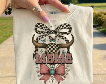Puede incluir: Sudadera de color crema con un diseño gráfico de una calavera a cuadros con cuernos largos, un lazo rosa y la palabra "MAMA" en letras rosas. El diseño incluye mariposas y detalles florales. La palabra "COMFORT" está impresa en el cuello.