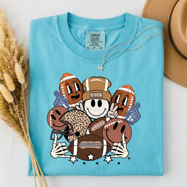 May include: A light blue t-shirt featuring a graphic of smiling footballs, a football helmet, and a smiley face wearing a football helmet. The word "FOOTBALL" is on the helmet. The shirt also has a Comfort Colors tag.