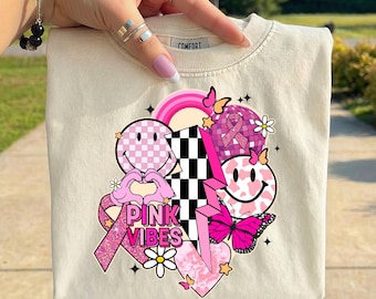May include: Cream-colored sweatshirt with a colorful graphic design. The design includes a pink ribbon, smiley faces, butterflies, a rainbow, and the words "PINK VIBES". The sweatshirt has a crew neck and the word "COMFORT" is printed on the collar.