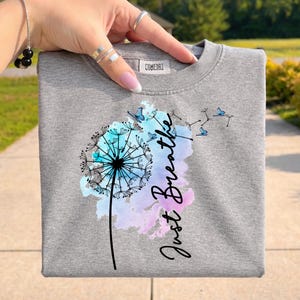 May include: A gray sweatshirt featuring a dandelion design with the words "Just Breathe." The dandelion is in shades of blue and teal, with butterflies. The text is in a cursive font. The sweatshirt has a crew neck.