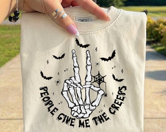 May include: A cream-colored t-shirt featuring a black graphic of a skeleton hand making a peace sign. The design includes small bats, spiders, and a spiderweb, with the text "PEOPLE GIVE ME THE CREEPS" arched around the bottom of the hand illustration.