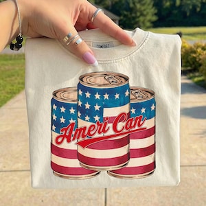 May include: Cream-colored t-shirt featuring three stylized beverage cans with a patriotic design. The cans have a blue top with white stars, red and white stripes, and the text "Ameri Can" in red script. The shirt has a crew neck.