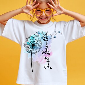 May include: White t-shirt featuring a dandelion design with blue butterflies and the words "Just Breathe" in a script font. The dandelion has a watercolor effect with blue and purple hues.